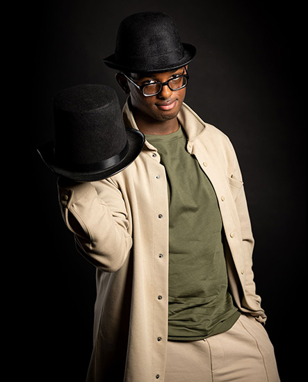 Mahdi - Digital Production, Design and Development Uxbridge College student - he is wearing glasses, a black bowler hat, green-t-shirt with a beige shirt and holding another black hat in his hand.