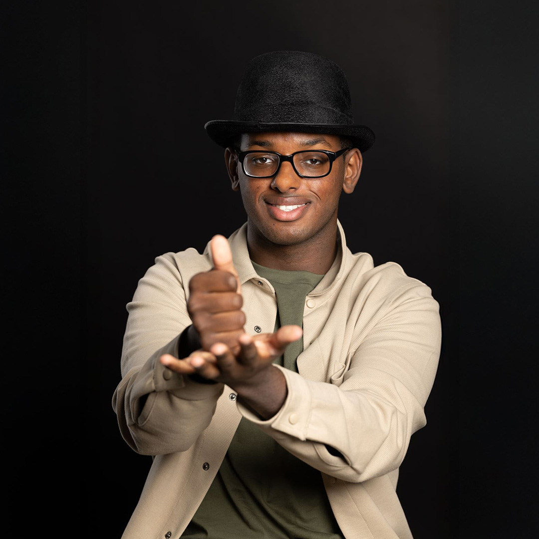 Mahdi - Digital Production, Design and Development Uxbridge College student - he is signing (BSL) and wears glasses, a black bowler hat, green-t-shirt with a beige shirt.