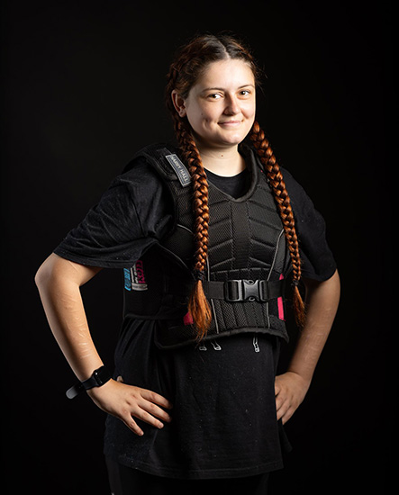 Alice - Uxbridge College Health and Social Care student - wearing a black vest with waist strap. She has her hands on her hips, red pigtails and is smiling.