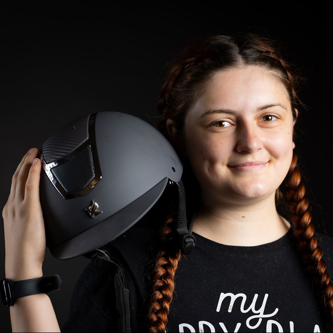 Alice - Uxbridge College Health and Social Care student - is holding a grey helmet under her shoulder. She has long red plaits and a black t-shirt on with white writing.