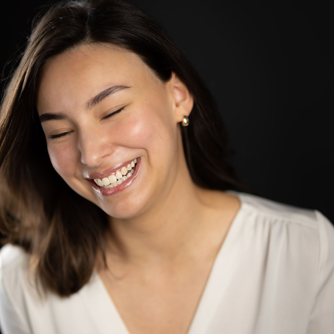 Anastasiia - Uxbridge College HND Business student - laughing with her eyes closed. She is wearing a white blouse and gold earrings. 