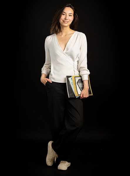 Anastasiia - Uxbridge College HND Business student - standing with one leg partly crossed over the other wearing a white blouse, black trousers and white sneakers. She is holding books in her left hand.