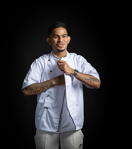 Johan - Uxbridge College Professional Cookery and Hospitality student - putting on his white chef's jacket and in mid smile. He has tattoos on both arms.