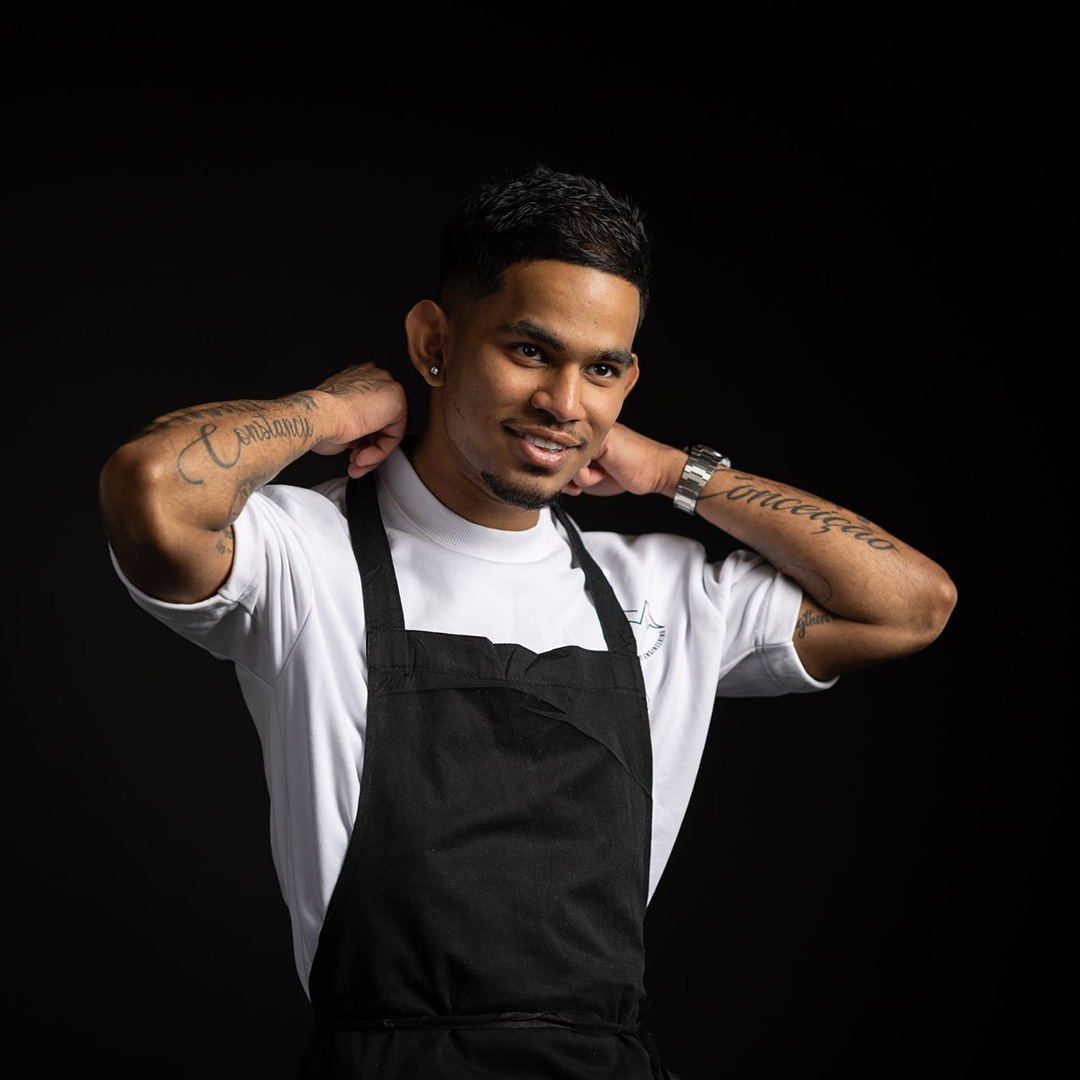 Johan - Uxbridge College Professional Cookery and Hospitality student - is adjusting his black apron from around his neck. He is wearing a white top and has tattoos on both arms.
