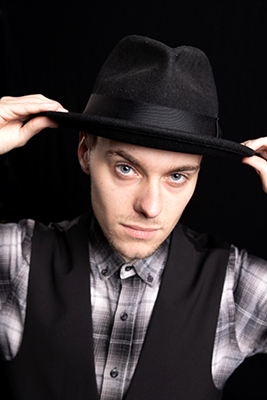 Stuart holds his hat. He wears a waistcoat and checked shirt.
