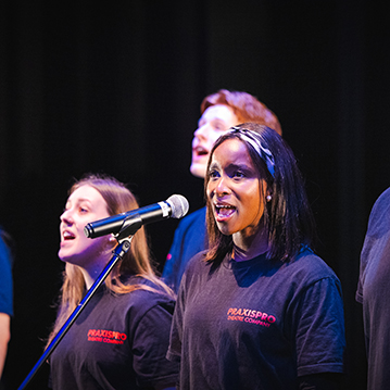 Praxis Performing Arts Students singing at the launch of the innovation partnership with MIT and HRUC. Students are singing on stage in front of a microphone all wearing black t-shirts. 