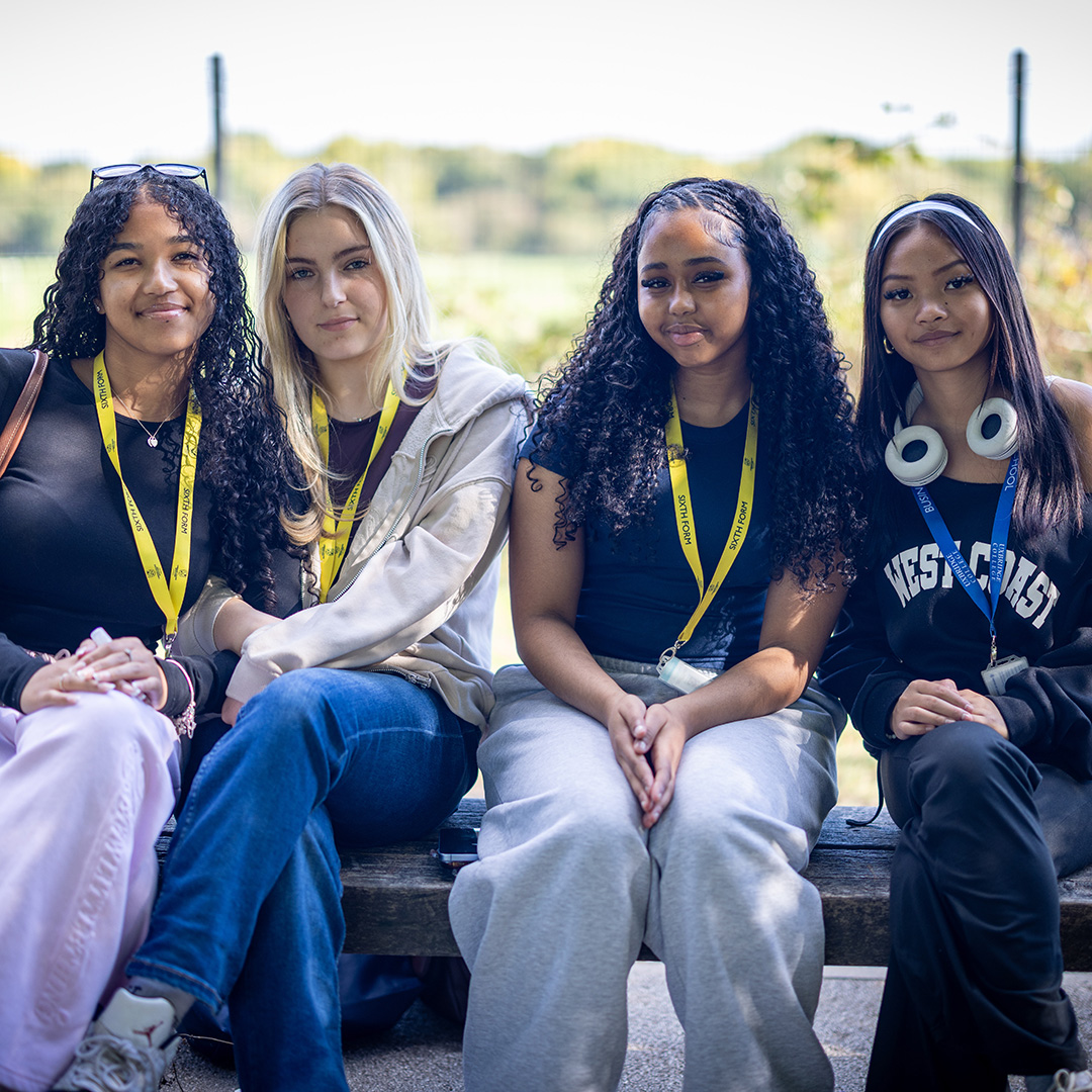 4 female Uxbridge Students sitting outside on a bench. They are wearing yellow and blue lanyards and there is grass and bushes behind them. 