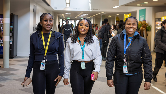 Three Uxbridge College students walking down the busy mall on the Uxbridge campus. There are other students and staff behind them. They are all wearing lanyards.