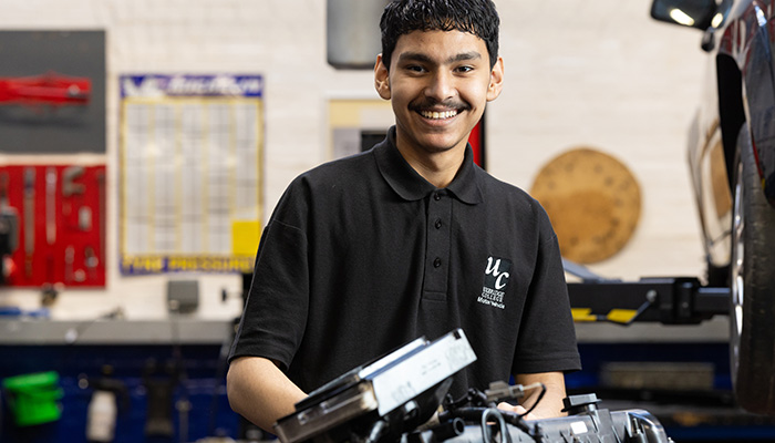 A male Uxbridge College Motor Vehicle student in the workshop. He is smiling wearing a black polo short with the Uxbridge College logo on it and there is an engine in front of him and a car on a car lift to the right.