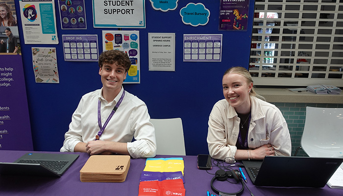 Two Uxbridge College Student Support staff at their booth at an Uxbridge Open Day. There are blue stand-up screens with information stuck to them. On the table in front of them are laptops, mouse pads, head phones, pens, buttons and travel card holders.