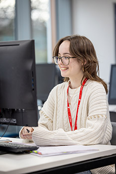Female student sitting in front of a keyboard and computer screen. She has long brown hair, wears glasses, a white jumper, red lanyard and is smiling.