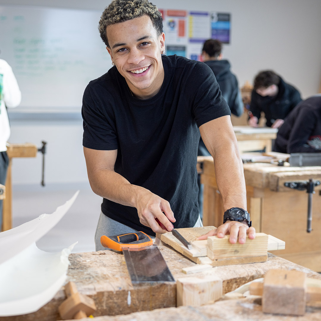 Male Richmond upon Thames College student in a workshop classroom with other students in the background, all working on wooden projects on benches. He is smiling and wearing a black t-shirt.