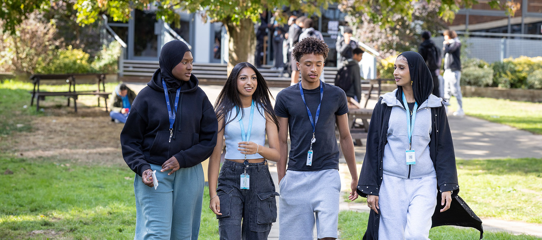 Four Uxbridge College students wallking in the outside garden area at the Uxbridge campus. They are all wearing black, grey or light blue clothing. 