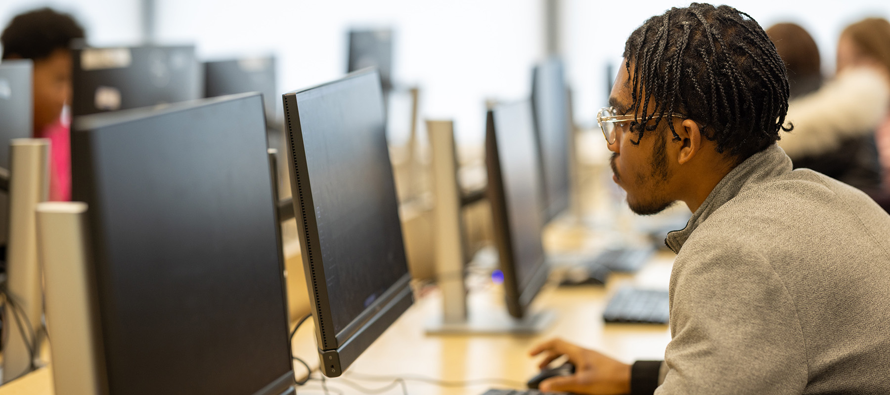 A male Uxbridge College student working at a computer. He is moving a computer mouse in his right hand and is wearing glasses. 