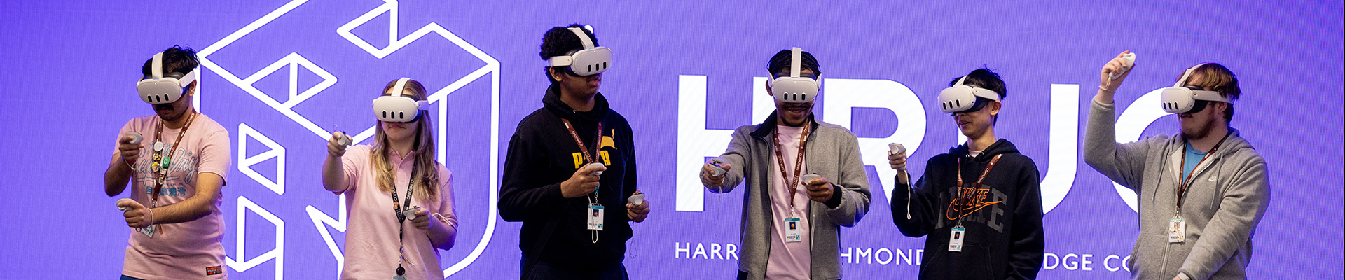Uxbridge College students all wearing VR headsets standing in front of a large screen with the HRUC logo on it and background in purple. 