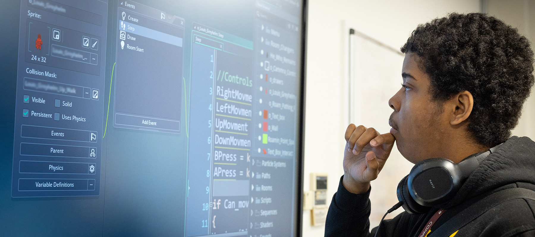 A male Uxbridge College computing student working on the electronic smart/blackboard. He has his hand up to his face and looks like he is thinking. He is wearing headphones around his neck. 