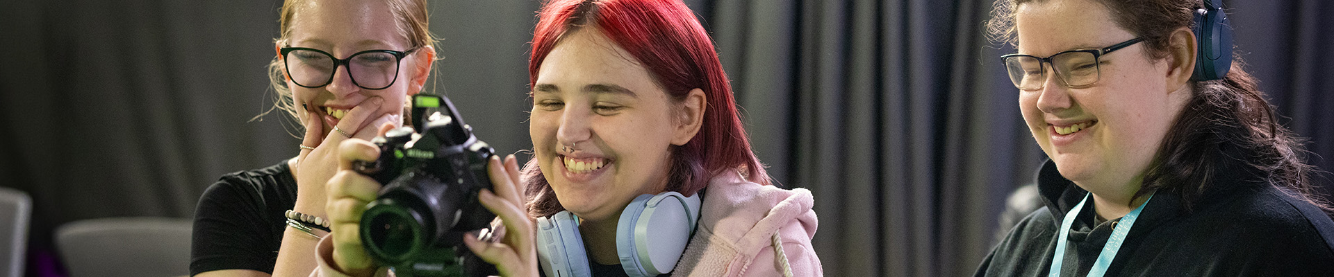 Three female Uxbridge media students using a camera on a tripod to take a photo and laughing. Two of them are wearing headphones and two are wearing glasses, the middle student has red hair.