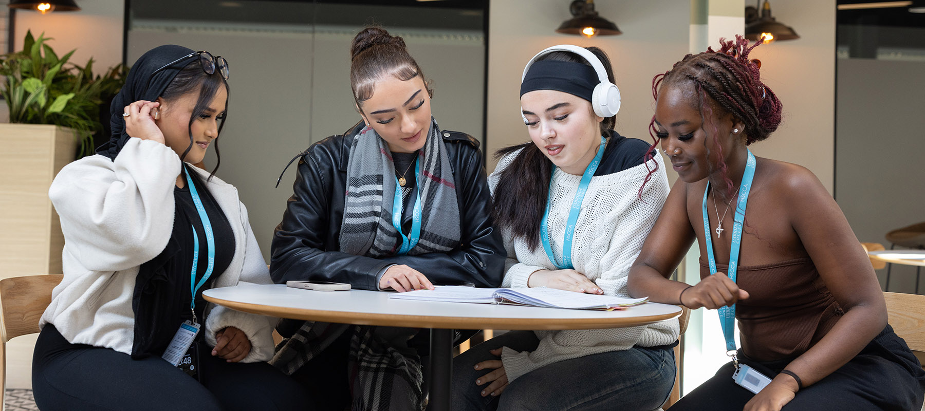 A group of female Uxbridge College students sitting around a table in the mall area. They have a workbook open and are all looking at it.