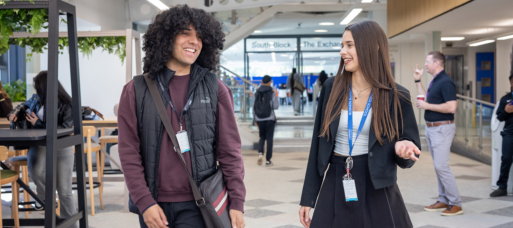An Uxbridge College male and female student talking and walking down the mall. There are other students and staff in the background. 