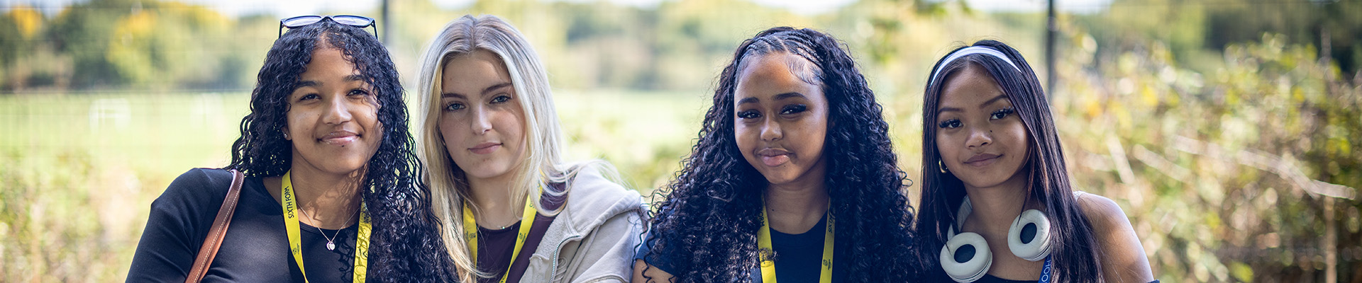 4 female Uxbridge Students sitting outside on a bench. They are wearing yellow and blue lanyards and there is grass and bushes behind them. 
