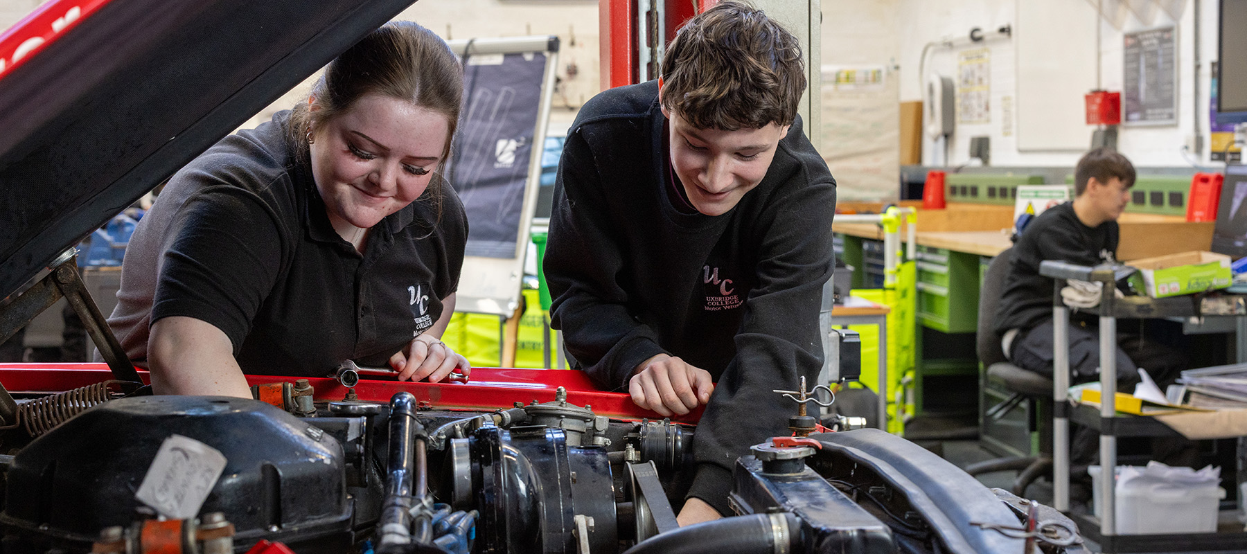 Male and female Uxbridge College Motor Vehicle students working on an engine under a red car bonnet. They are in the workshop with other students behind them. 