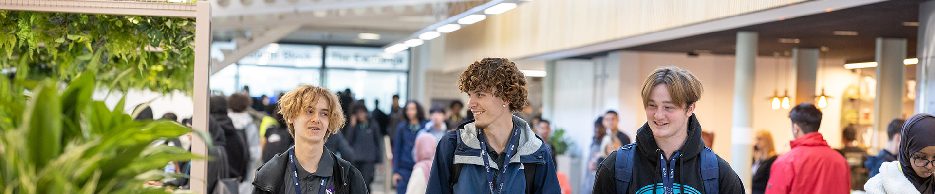 3 male Uxbridge College students walking down the modernised mall at the Uxbridge campus. Smiling at each other and all wearing lanyards. There are many students in the background.  