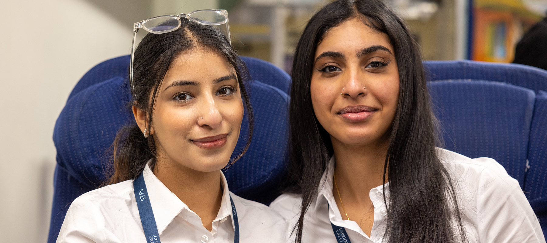 2 female Uxbridge Travel and Tourism students smiling at the camera. They are wearing white shirts and sitting in blue chairs from the mock aircraft cabin.