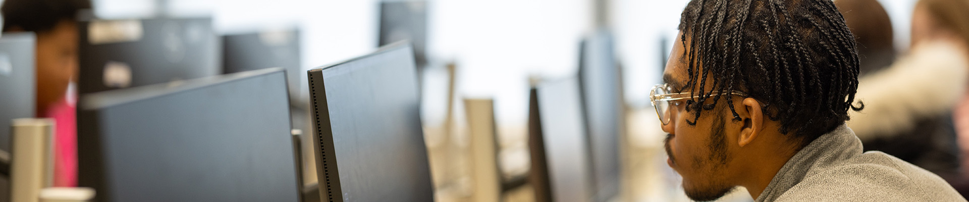 A male Uxbridge College student sitting at a row of computers with a computer screen and keyboard in front of him. His hand is on a mouse and he is concentrating on what is on the screen. He is wearing glasses and a grey jacket. 