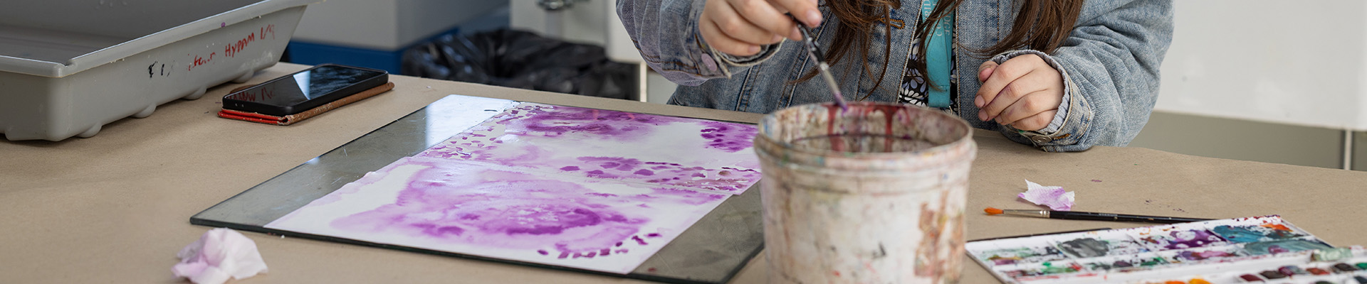 A female Uxbridge College student working in the arts studio on a project. She is painting a surface with purple paint and there is a bucket she is dipping her paint brush into in front of her with a paint palette next to it. She is wearing black headphones over her ears, has red glassses and long brown hair. 