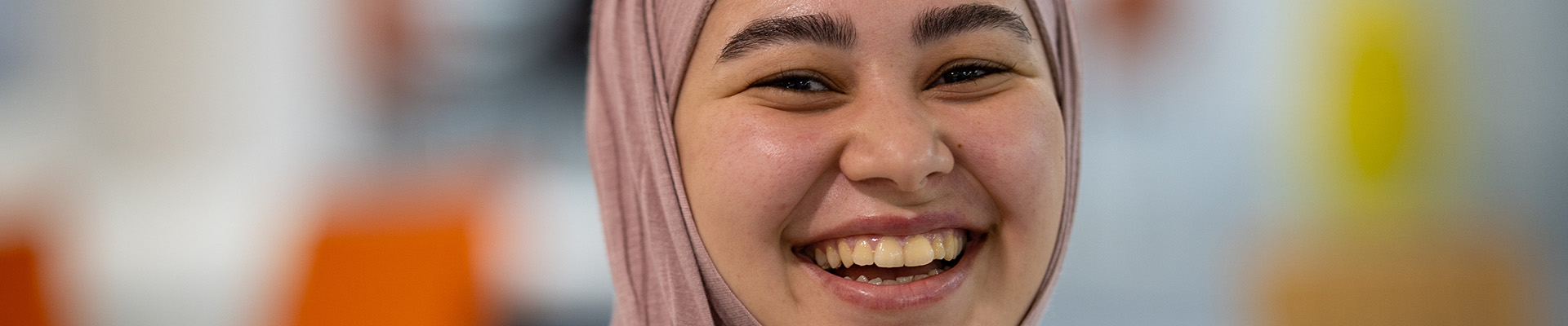 Female ESOL Uxbridge College student smiling. She is wearing a pink headscarf, yellow lanyard and a colourful pastel top.