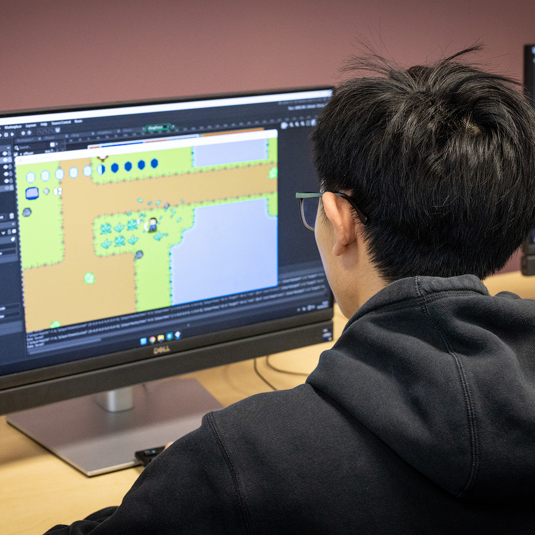 A male Uxbridge College computing games development student working on a games project. He is looking at a computer screen and there is a picture of a game on there.