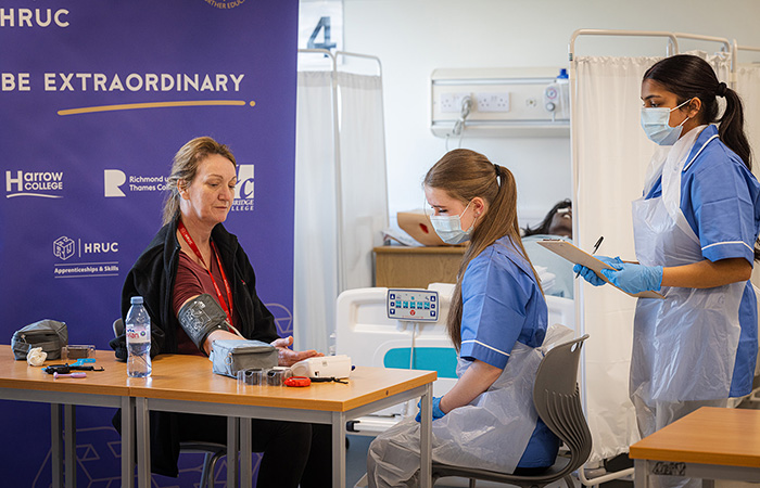 Two female Uxbridge College Health and Social Care students practising taking blood pressurre from a woman. The students are wearing protective PPE masks and aprons over their blue uniforms. There is a hospital bed behind them.