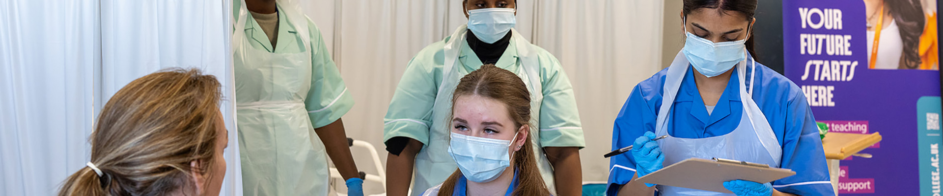 Uxbridge College Health and Social Care students practising taking blood pressurre. They are all wearing protective PPE masks and aprons over their uniforms.