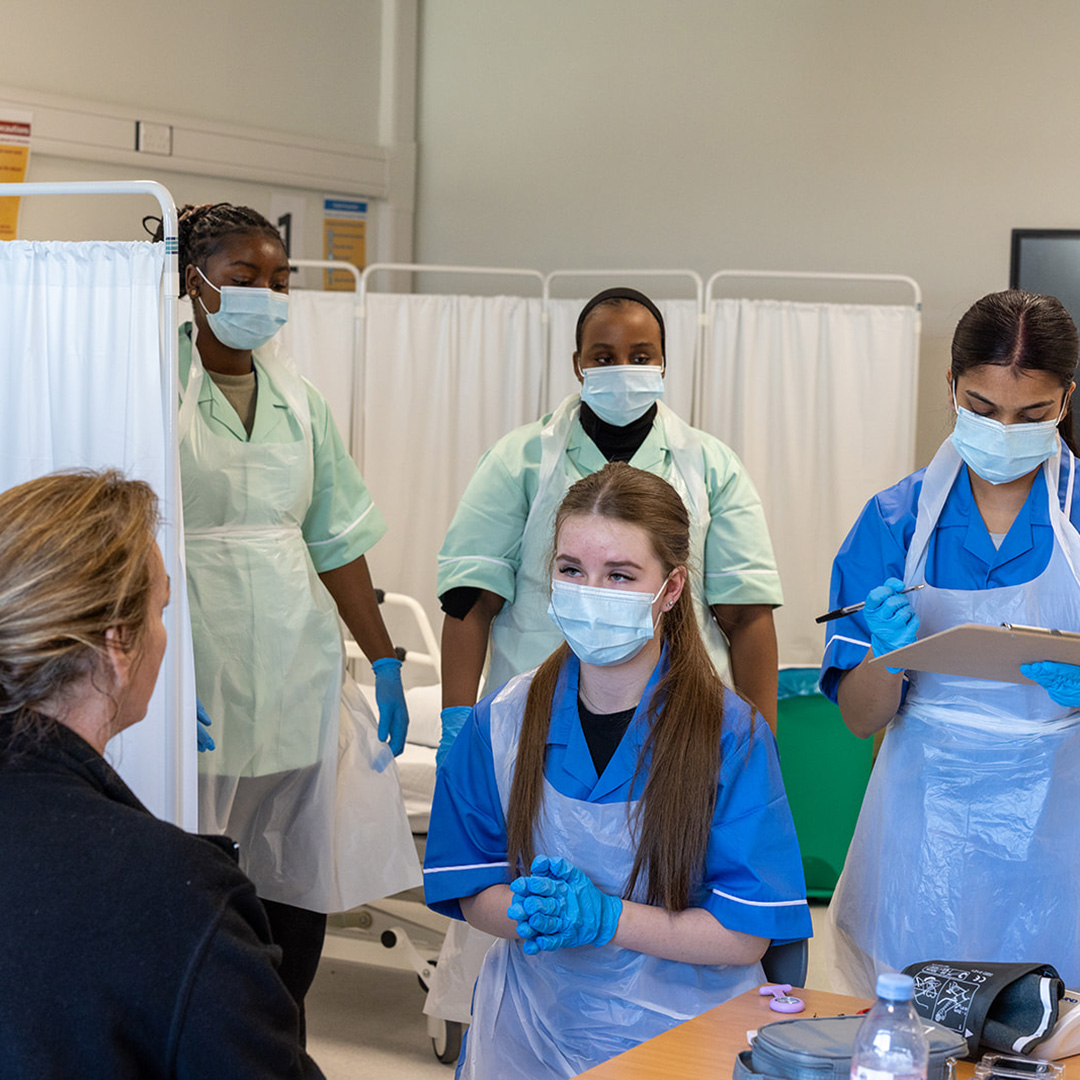 Uxbridge College Health and Social Care students practising taking blood pressurre. They are all wearing protective PPE masks and aprons over their uniforms.