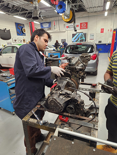 Haider, Uxbridge College Motor Vehicle Maintenance student, working on an engine in the Uxbridge workshop. He is wearing a blue coverall and white gloves.