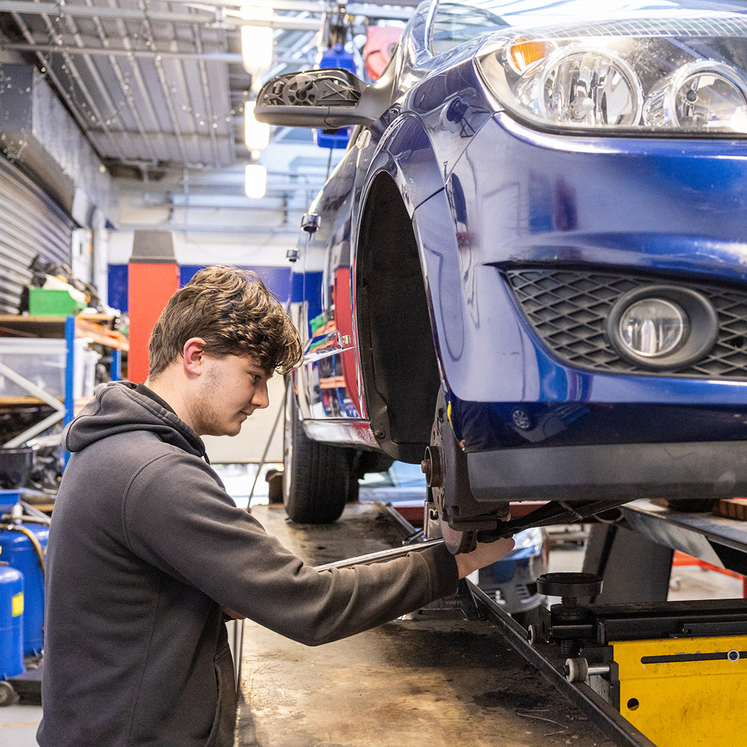 Male Uxbridge College student working on a car. The car is blue and hoisted up so he can work on the parts underneath.