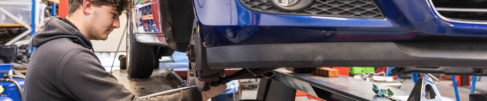 Male Uxbridge College student working on a car. The car is blue and hoisted up so he can work on the parts underneath.