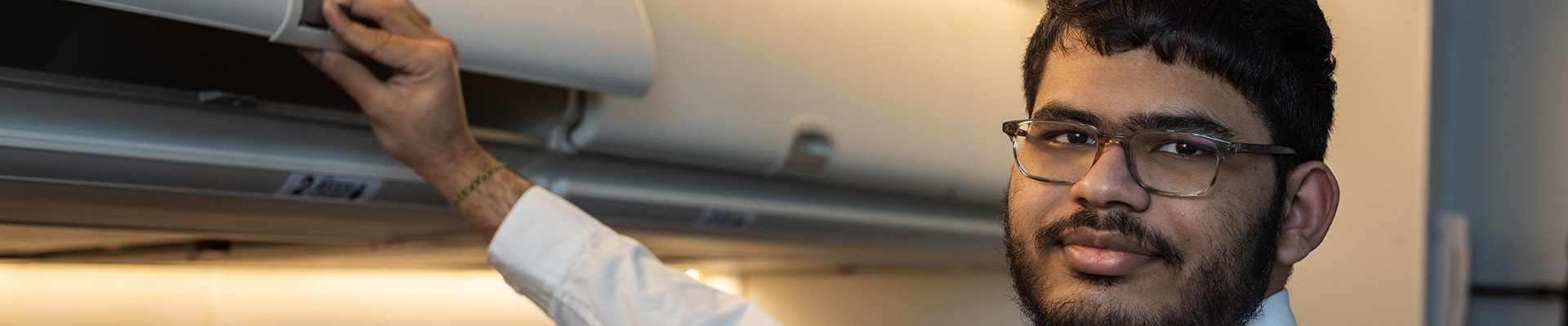 A male Uxbridge College Aviation, Travel and Tourism student in the mock cabin closing the overhead compartments as part of the onboarding routine for an aircraft. He has a beard and glasses and is wearing a white shirt and pale blue tie. Passangers are in the seats behind him. 