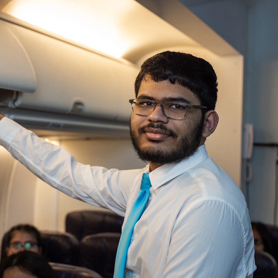 A male Uxbridge College Aviation, Travel and Tourism student in the mock cabin closing the overhead compartments as part of the onboarding routine for an aircraft. He has a beard and glasses and is wearing a white shirt and pale blue tie. Passangers are in the seats behind him. 
