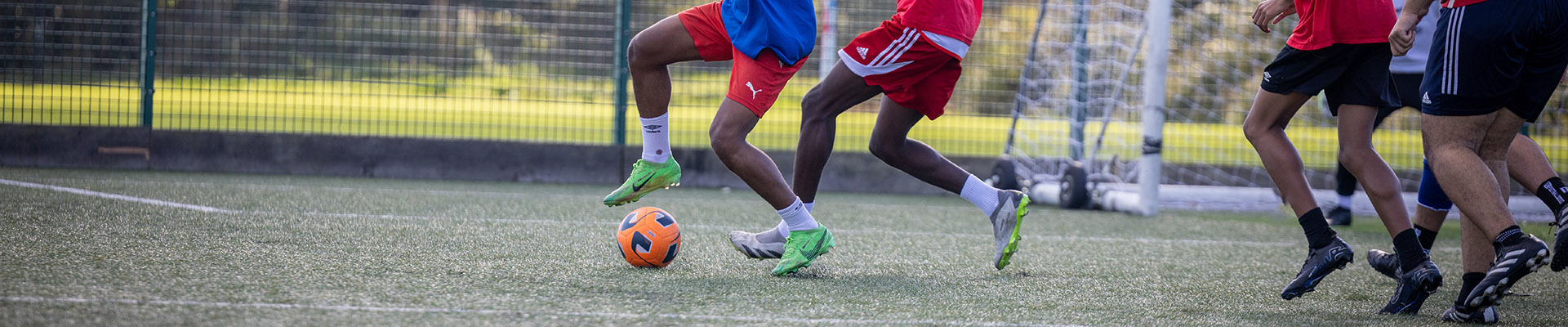 Uxbridge College students playing football on an outside pitch. They are running to catch the bright orange ball.