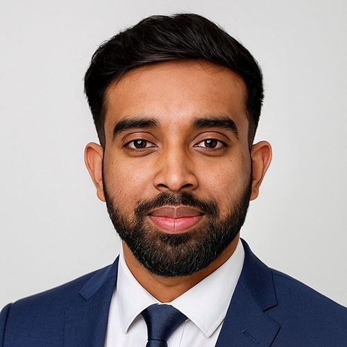 Osric (international student) is smiling at the camera, wearing a blue suit and tie with a white shirt, he also has a beard. 