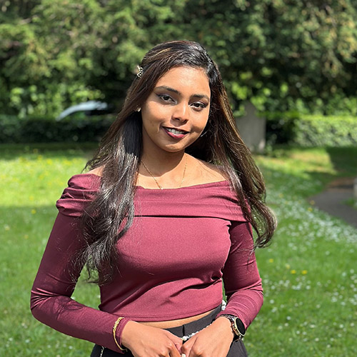 Venica wearing a red long sleeve top, off the shoulders, with a green garden landscape behind her. 