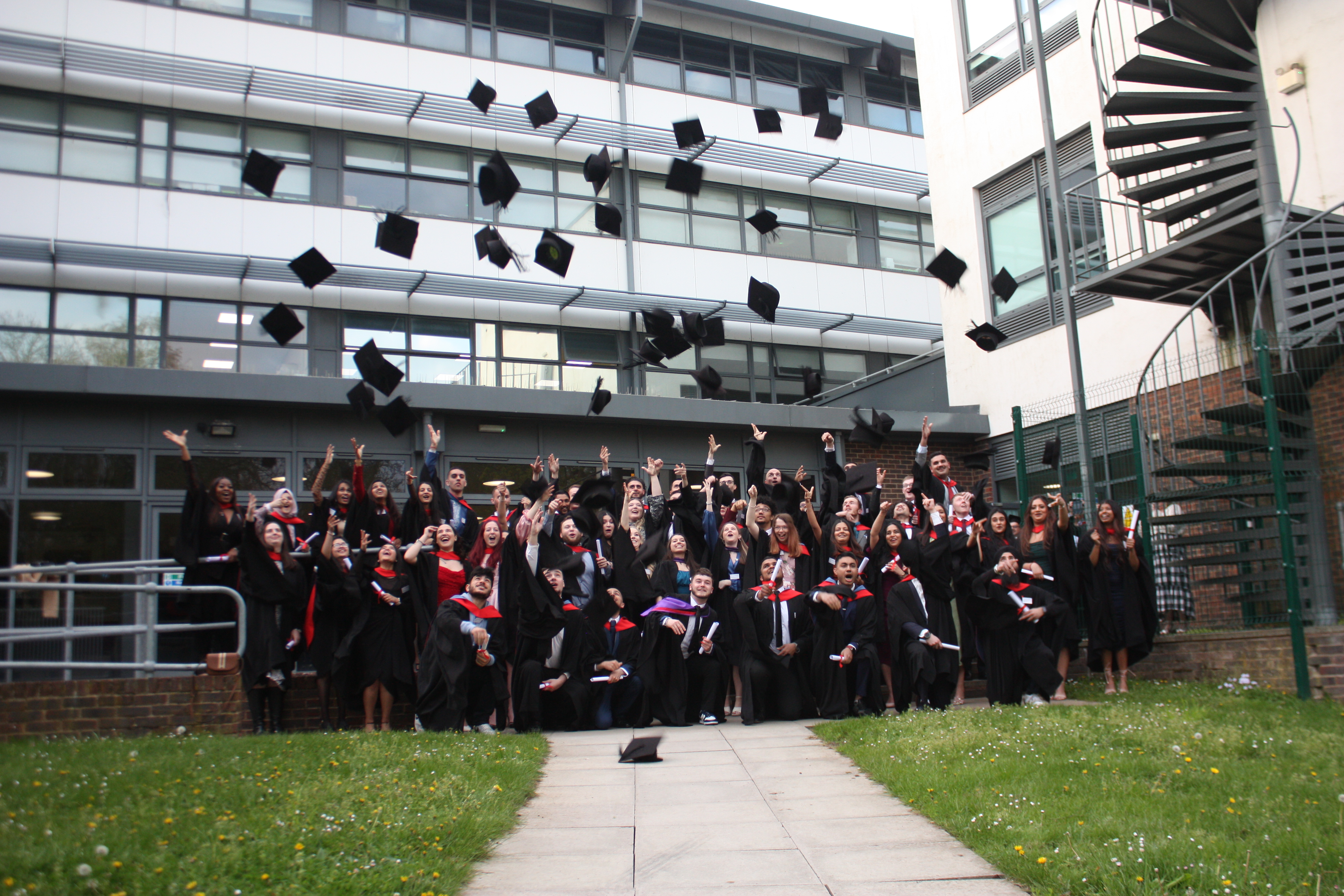 Group of 50 Uxbridge College and Harrow College graduates outside the Uxbridge College building throwing caps in the air at graduation ceremony