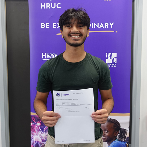 A male Uxbridge student showing his A-Level results and smiling broadly in front of a banner that has the HRUC logo and a purple background.     