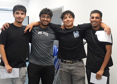 A group of 4 male Uxbridge students with their arms across their shoulders, smiling and holding their letters of results. They are all wearing black and grey.         