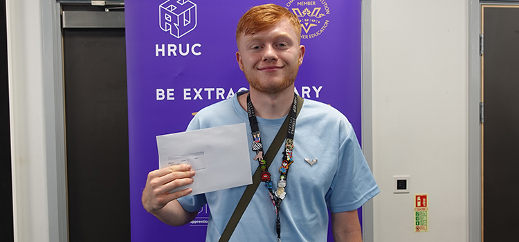 A male Uxbridge student showing his results envelope and smiling in front of a banner that has the HRUC logo and a purple background. 