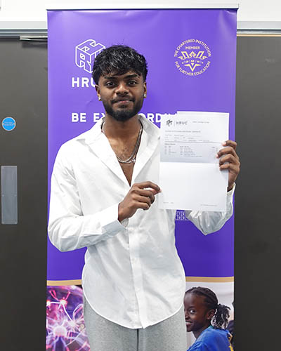 Male Uxbridge College student holding up his GCSE results letter proudly and smiling at the camera. He is standing in front of a purple Be Extraordinary banner with the HRUC logo on it.                 