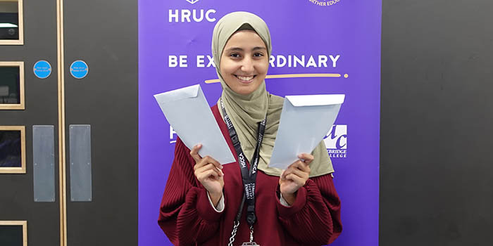 Female Uxbridge student holding her GCSE results envelopes. She is smiling at the camera and standing in front of a Be Extraordinary purple banner with HRUC logo.