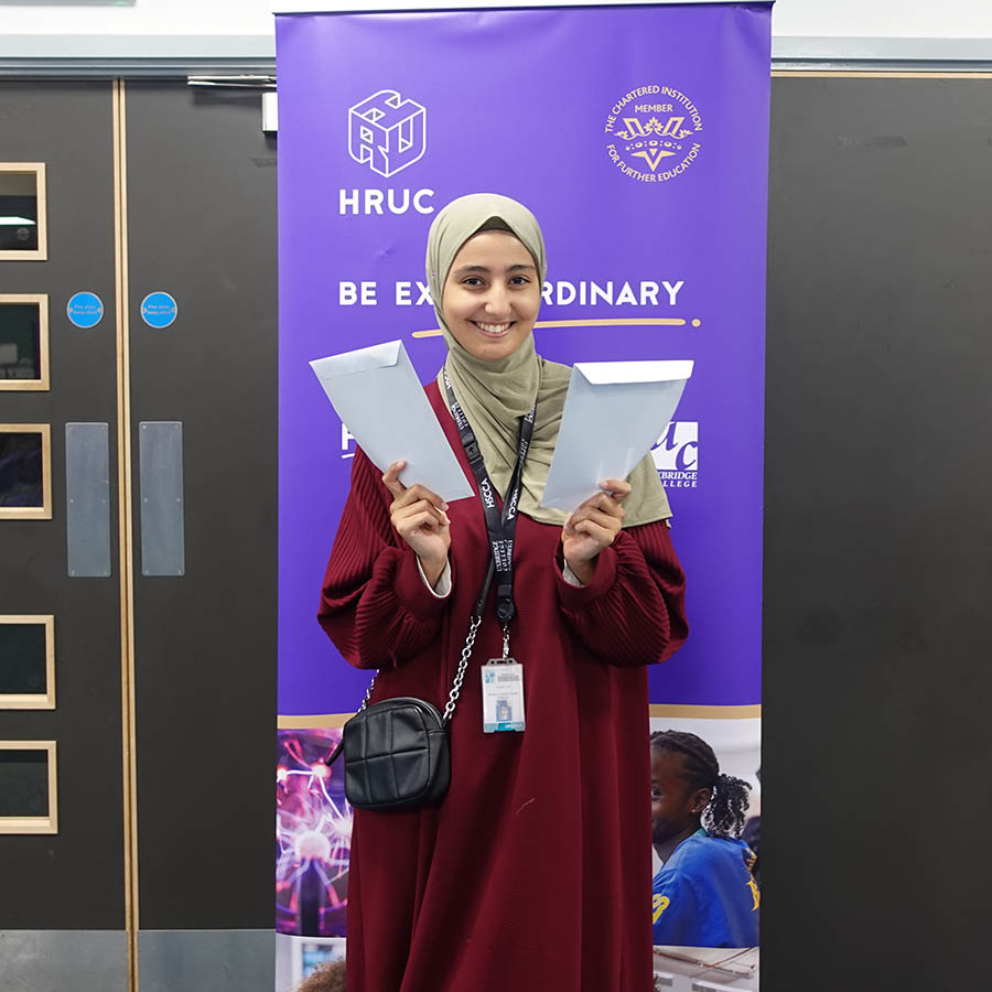 Female Uxbridge student holding her GCSE results envelopes. She is smiling at the camera and standing in front of a Be Extraordinary purple banner with HRUC logo.