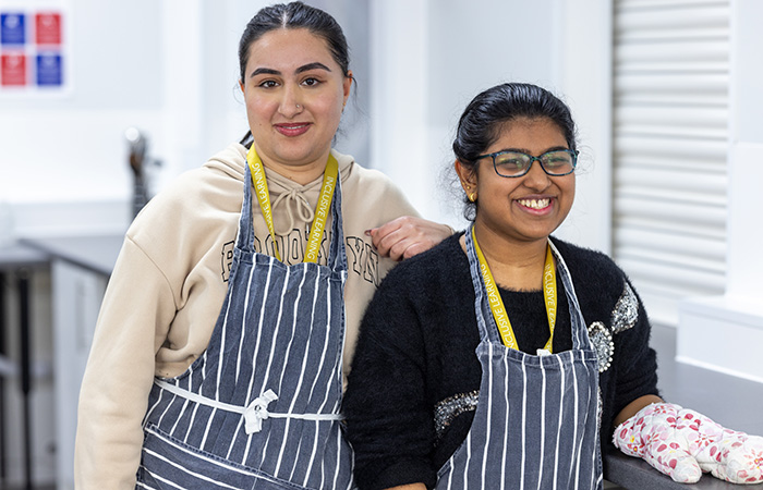 Two Female Uxbridge College students from Inclusive Learning in the kitchen at Hayes Campus. They are both wearing blue with white stripe aprons with yellow lanyards. 